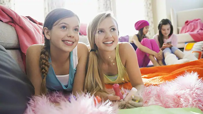 quatro meninas dentro de um quarto interagindo. Na frente, são duas meninas de cabelos claros, uma com tranças e vestindo blusa azul com sobreposição branca e a outra com cabelo preso e blusa amarela. Atrás, duas meninas de cabelos escuros, vestindo roupas rosas, uma delas usando capuz e sentadas olhando para o celular.