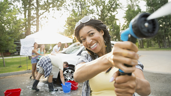 Uma mulher negra com cabelos longos nos ombros, vestindo blusinha amarela, está sorrindo enquanto aponta uma mangueira que esguicha água. Ao fundo há cinco pessoas lavando um carro branco.