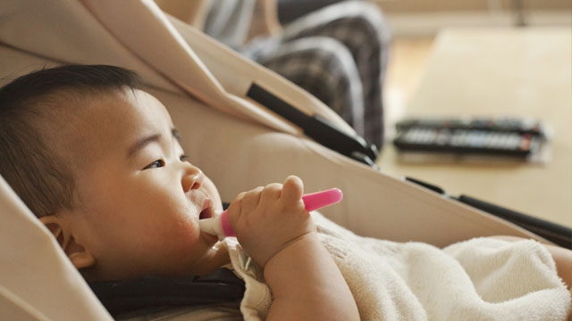 bebê deitado em um bebê conforto e segurando em uma das mãos uma escova de dentes que está na boca.