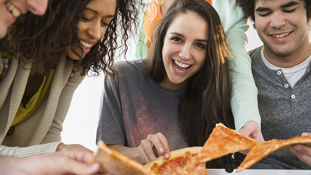 grupo de quatro amigos sorridentes comendo uma pizza. Na ponta está o perfil de um homem, no meio uma mulher negra com blusa verde casaco bege, ao seu lado uma moça branca de cabelos longos e escuros, vestindo uma camiseta cinza-escuro com efeito sombreado e na outra ponta um rapaz de cabelos crespos escuros, camiseta branca e blusa cinza