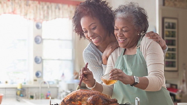 duas mulheres negras com cabelos curtos, uma é uma senhora de idade avançada com cabelos grisalhos e está vestindo um avental verde água enquanto tempera um chester, a outra mulher está abraçando a senhora e sorri enquanto olha para o alimento.