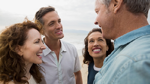 quatro pessoas sorrindo: à direita um homem com cabelos grisalhos vestindo camisa azul social. Ao seu lado, uma mulher com cabelos longos e ondulados veste uma blusinha azul escura com gola branca. No lado dela, um homem com cabelos curtos e claros veste camisa social esverdeada de manga curta. À esquerda uma mulher tem cabelos longos e ondulados.