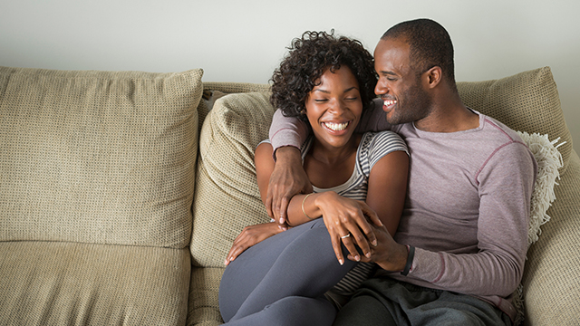 Casal. Homem e mulher, negros, com idade aproximada de 35 anos, sentados no sofá se abraçando e sorrrindo. Ele tem o cabelo bem curto, quase raspado, veste camisa rosa clara acinzentada e calça cinza escura. Ela tem cabelo castanho escuro cacheado, veste blusinha de manga curta, listrada, cinza e branca, e veste calça leggin cinza escura. 