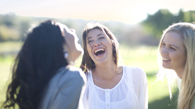 três amigas, uma loira, a outra de cabelos castanhos e camisa clara e outra morena com blusa clara em um ambiente cercado de natureza, todas sorrindo