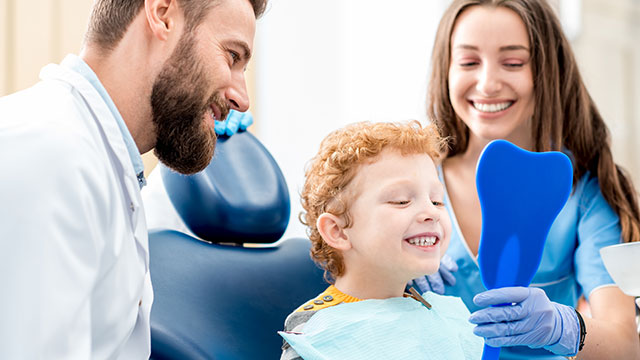 um menino sentado em uma cadeira de consultório odontológico olhando para um espelho em formato de dente na cor azul, ele tem cabelos encaracolados e ruivos, ao lado esquerdo dele tem uma mulher adulta com cabelos longos e escuros, vestindo jaleco azul e luvas, ao lado direito tem um homem com cabelos curtos e barba, vestindo camisa social azul.