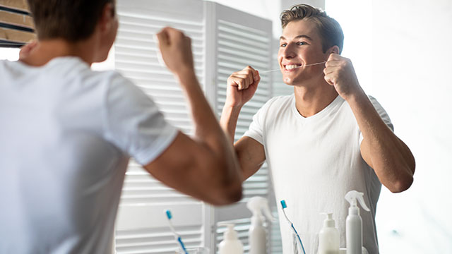 homem jovem, com cabelos loiros, vestindo uma camiseta branca, passando fio dental nos dentes em frente a um espelho