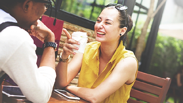 Mulher e homem em um restaurante. A mulher, que aparece de frente, está sorrindo. O homem, que está de costas na foto, é negro, está usando uma camisa social branca e um suspensório preto. 