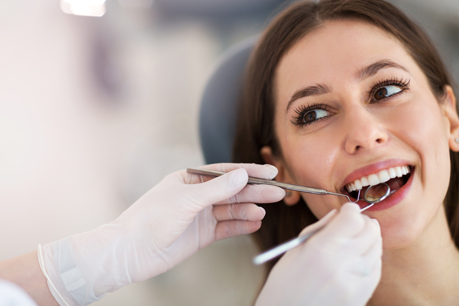 Mulher jovem de cabelos escuros, com a boca aberta e sorrindo levemente enquanto o dentista examina sua boca com o auxílio de instrumentos. Apenas as mãos do dentista estão à mostra, com luvas.