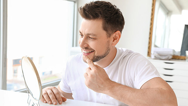 Homem branco no quarto sorrindo para um espelho de mesa 